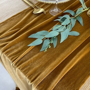 Könnte beinhalten: Ein goldfarbener Samttischläufer mit einem Zweig grüner Blätter darauf. Der Läufer ist an einigen Stellen gefaltet, wodurch ein strukturierter Look entsteht.