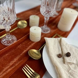 Puede incluir: Un camino de mesa de terciopelo marrón con dos velas blancas, dos cucharas doradas, un tenedor dorado, dos copas de vino transparentes y una servilleta de lino blanco con dos pequeñas piñas en un plato blanco.