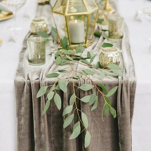 May include: A gray velvet table runner with a sprig of eucalyptus leaves and gold candle holders. The runner is on a white tablecloth.