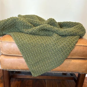 May include: A chunky knit throw blanket in a solid olive green color. The blanket features a dense, textured stitch pattern with a visible loop structure throughout the fabric, creating a thick and tactile surface that is draped over a piece of furniture.