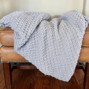 May include: A light gray knitted blanket draped over a brown leather ottoman. The blanket has a textured appearance and is folded to show its thickness and weave. The neutral color and soft texture suggest comfort and warmth.