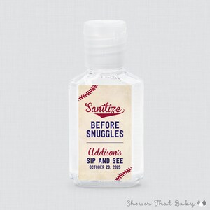 May include: Clear plastic hand sanitizer bottle with a white cap. The label reads "Sanitize BEFORE SNUGGLES" in red and blue, with "Addison's SIP AND SEE OCTOBER 20, 2025" below. Baseball stitching accents the design.