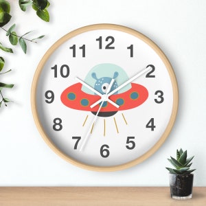 May include: A white clock with a wooden frame features a red and blue cartoon spaceship with a black eye. The clock face has black numbers 1 through 12.