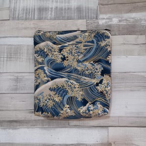 May include: A rectangular zipped pouch featuring a repeating pattern of stylised ocean waves in shades of deep navy blue, light blue, and cream. The design includes intricate line work and foam details, finished with a black zip at the top.