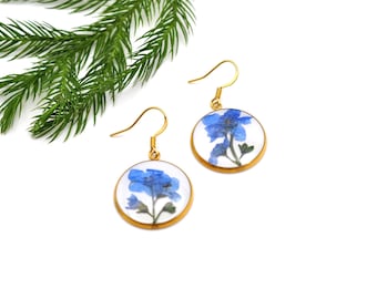May include: A pair of gold-colored, round earrings with a pressed blue flower design set against a white background. The earrings have a hook closure. Green foliage is in the background.
