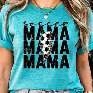 Peut inclure: T-shirt turquoise avec l'inscription "soccer mama" en écriture noire au-dessus de trois lignes superposées de "MAMA" en gras, police vieillie. Un ballon de foot et un éclair sont au centre du t-shirt.