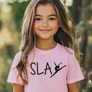 May include: A light pink t-shirt with the word "SLAY" in black script and a black silhouette of a dancer performing a split leap. The girl is smiling and has long brown hair.