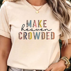 May include: A beige t-shirt with the phrase "MAKE heaven CROWDED" printed across the front in a mix of fonts and colors. The word "MAKE" is in pink and leopard print, "heaven" is in black script, and "CROWDED" is in various colors and leopard print.