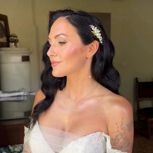 以下が含まれることがあります： 黒髪の女性が、オフショルダーの白いドレス、金のネックレス、パールのイヤリングを身に着けています。パールが付いた金のヘアクリップが髪を飾っています。女性はメイクをしており、腕にタトゥーがあります。画像は屋内で撮影されています。