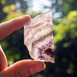 May include: A hand holding a piece of amethyst with purple and white banding. The amethyst is translucent and has a natural, rough texture.