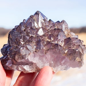 Könnte beinhalten: Ein Haufen Amethystkristalle, gehalten in einer Hand, mit einem Feld und blauem Himmel im Hintergrund. Die Kristalle sind eine Mischung aus Lila und Klar, mit scharfen, spitzen Formationen. Der Gesamteindruck ist natürlich und erdig.