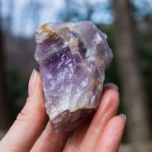 Könnte beinhalten: Ein großer, violetter Amethystkristall mit rauer, natürlicher Textur. Der Kristall hat eine tiefviolette Farbe mit einigen helleren Bereichen und einigen weißen Streifen.