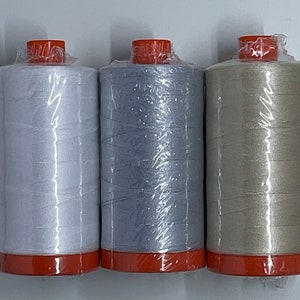 May include: Three spools of thread in white, gray, and beige, each wrapped in clear plastic. The spools have orange caps and bases. These are likely for sewing or embroidery projects.