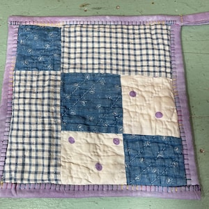May include: A patchwork pot holder with a blue and white checkered pattern, a blue floral pattern, and a white background with purple polka dots. The pot holder has a purple border and a purple loop handle.