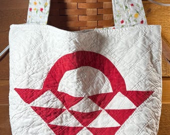 May include: A quilted tote bag featuring a white fabric base with a prominent red geometric basket-weave pattern. The bag includes two shoulder straps made from a white fabric decorated with a small, colorful floral print of red and yellow flowers.