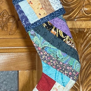 May include: A patchwork Christmas stocking with a red, blue, green, and white color scheme. The stocking is made of different fabric scraps sewn together in a diagonal pattern.