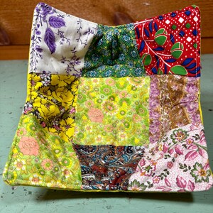 May include: A yellow fabric bowl with a patchwork design. The bowl is made of nine different squares of fabric, each with a different floral pattern. The fabric squares are sewn together to create a unique and colorful design.