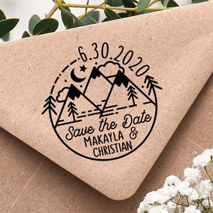 Könnte beinhalten: Ein brauner Papierumschlag mit einem schwarzen runden Stempel. Der Stempel zeigt Berge, Bäume, einen Mond und den Text "6.30.2020" und "Save the Date MAKAYLA & CHRISTIAN". Grüne Blätter und weiße Blumen umgeben den Umschlag.