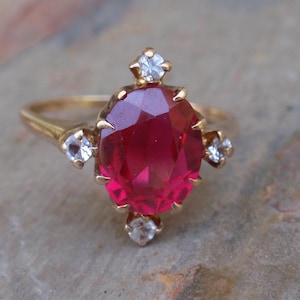 Peut inclure: Une bague en or avec une grande pierre précieuse rouge foncé de taille ovale au centre. La pierre centrale est entourée de quatre petites pierres d'accent claires de taille ronde, placées en haut, en bas, à gauche et à droite du sertissage.