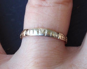 May include: A gold-toned ring with a decorative band design. The ring is on a finger, showcasing its intricate details and polished finish. The band features a series of rectangular and patterned elements.