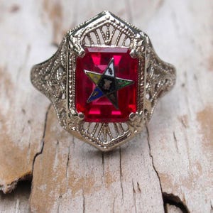 May include: A silver ring with a red gemstone set in a filigree design. The gemstone features a five-pointed star with a yellow, blue, and white design.