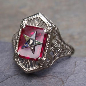May include: A silver filigree ring with a red gemstone set in the center. The gemstone features a five-pointed star with a smaller star inside.
