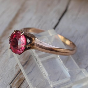 May include: A rose gold ring featuring a large, oval-cut, pink gemstone. The ring is set on a clear plastic display stand against a weathered wooden surface. The gemstone is the focal point, catching the light and showcasing its vibrant color.