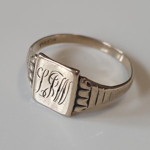May include: A silver signet ring with a square face featuring the engraved initials "LBM". The ring has a simple band and decorative detailing on the shoulders. The ring is likely vintage or antique.