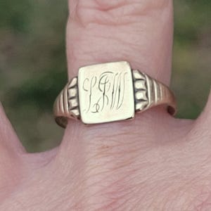 May include: A gold signet ring with a square face, engraved with the initials "LFW" in an elegant script. The ring has a classic design with a ribbed band and a polished finish, showcasing a vintage aesthetic.