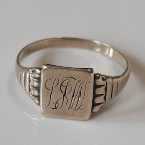 May include: A gold-toned signet ring with a square face and engraved initials. The ring has a simple band with ribbed detailing on the shoulders. The initials are in an elegant script. The ring is set against a white background.
