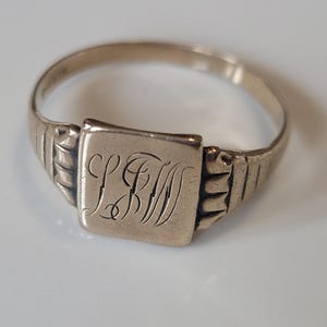 May include: A vintage gold-coloured signet ring. The ring has a square face with engraved initials. The band features a ribbed design on the sides. The ring is set against a white background.
