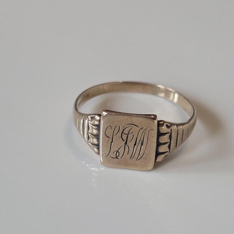 May include: A gold-toned signet ring with a square face and engraved initials. The ring has a simple band with ribbed detailing on the shoulders. The initials are in an elegant script. The ring is set against a white background.