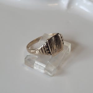 May include: A silver ring with a rectangular face engraved with initials. The ring has a plain band and decorative shoulders. The ring is displayed on a clear, rectangular stand.