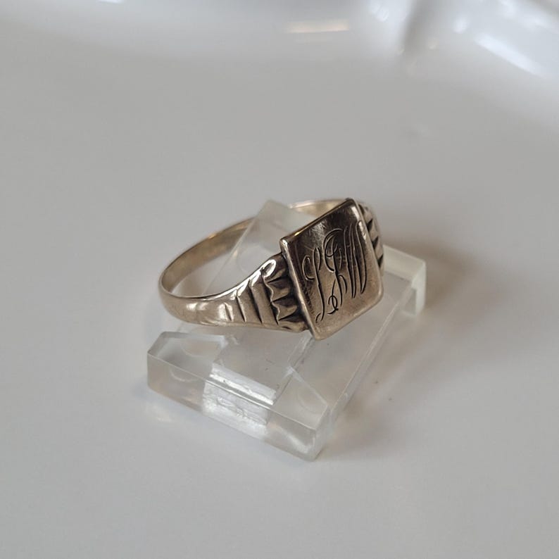 May include: A silver ring with a rectangular face engraved with initials. The ring has a plain band and decorative shoulders. The ring is displayed on a clear, rectangular stand.