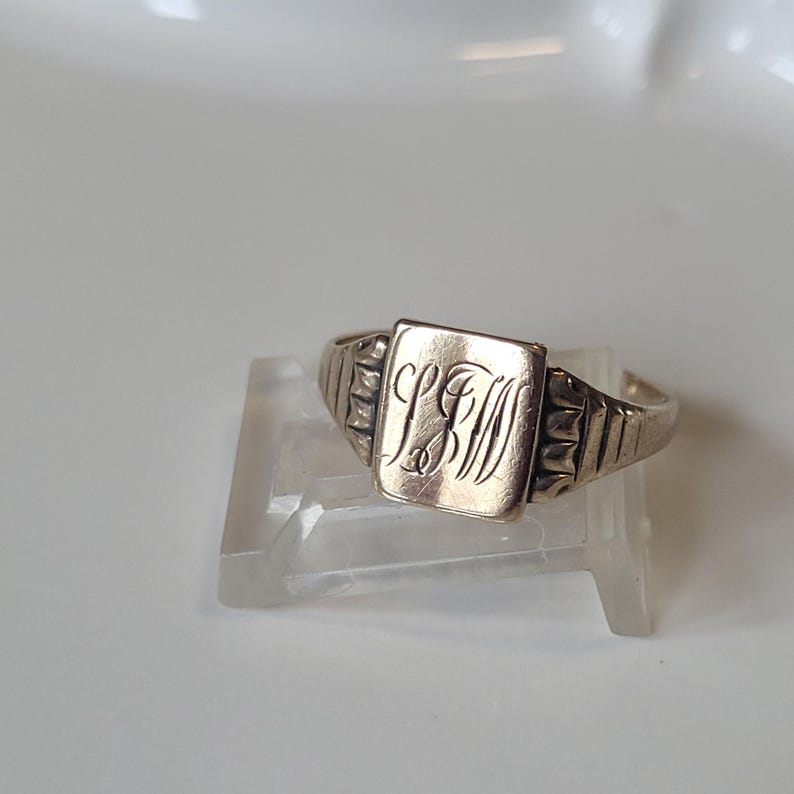 May include: A silver ring with a rectangular face engraved with initials. The band has a textured design on the sides. The ring is displayed on a clear stand against a white background.