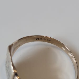 May include: Close-up of a gold ring with the inscription "OB 10K" on the inside band. The ring has a smooth, polished surface and a classic design. The ring is likely made of gold.