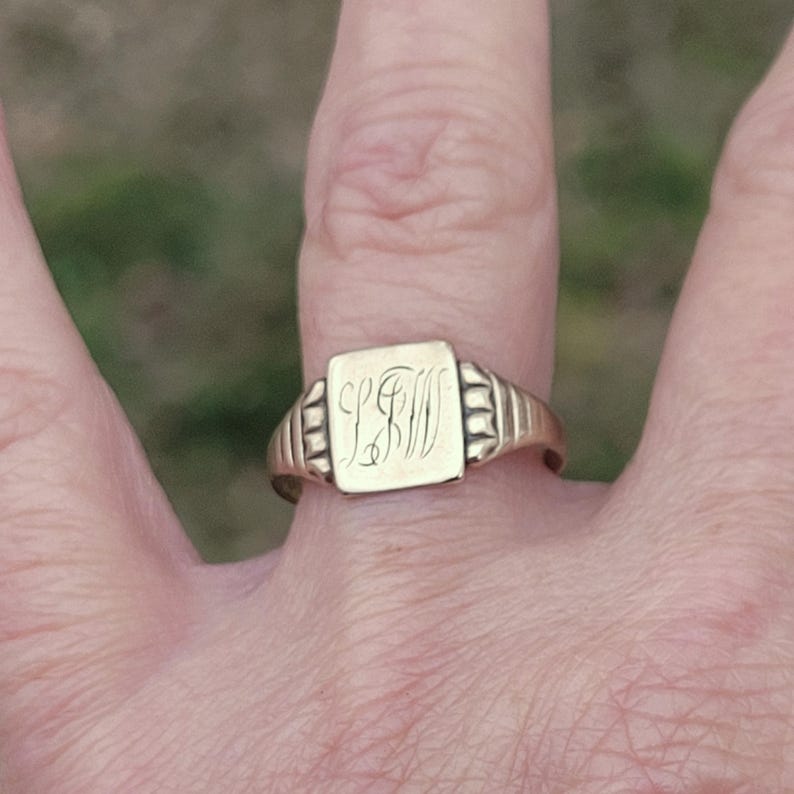 May include: A gold signet ring with a square face, engraved with the initials "LFW" in an elegant script. The ring has a classic design with a ribbed band and a polished finish, showcasing a vintage aesthetic.