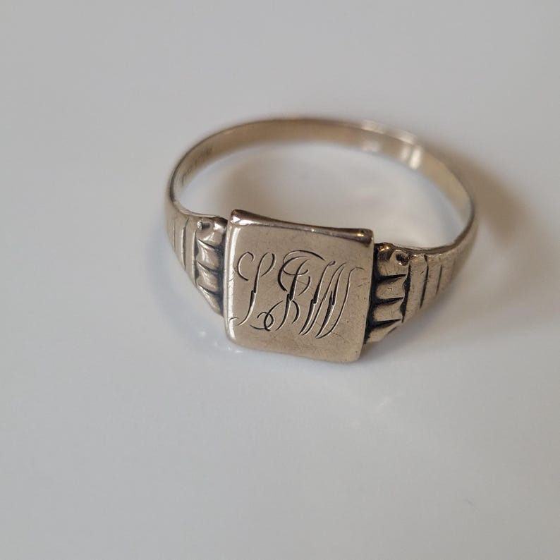 May include: A vintage gold-coloured signet ring. The ring has a square face with engraved initials. The band features a ribbed design on the sides. The ring is set against a white background.