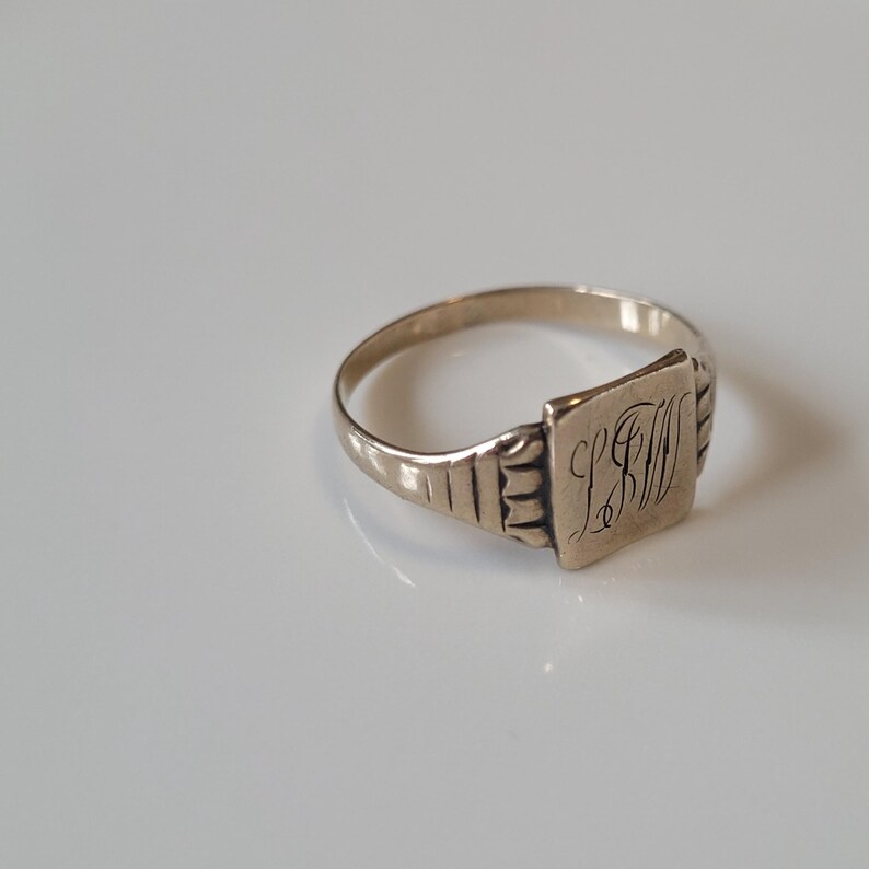 May include: A silver signet ring with a square face bearing engraved initials. The ring has a plain band and decorative shoulders. The initials are in a cursive script. The ring is set against a white background.