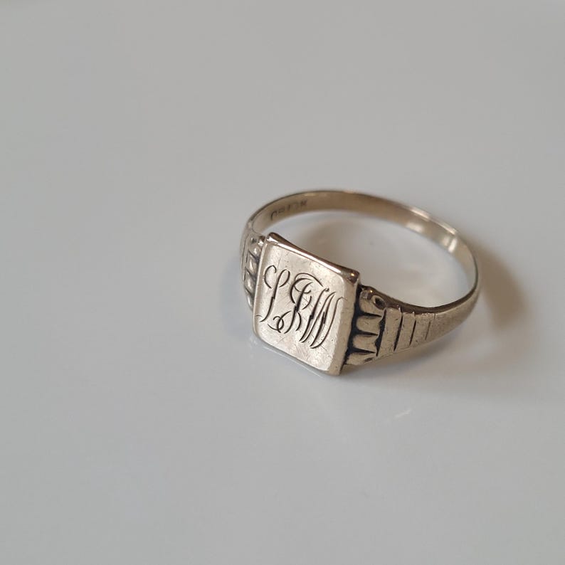 May include: A silver signet ring with a square face featuring the engraved initials "LBM". The ring has a simple band and decorative detailing on the shoulders. The ring is likely vintage or antique.