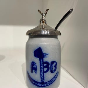 Puede incluir: Un tarro de cerámica blanco con un diseño de ancla azul y las letras "A B O" en la parte delantera. El tarro tiene una tapa plateada con una pequeña figurita plateada de una persona haciendo una parada de manos en la parte superior.