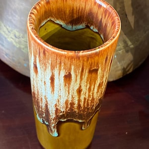 May include: A tall, cylindrical ceramic vase with a glossy finish. The vase features a color gradient, transitioning from a dark brown at the top to a mustard yellow at the base. The brown glaze drips down, revealing a turquoise hue.