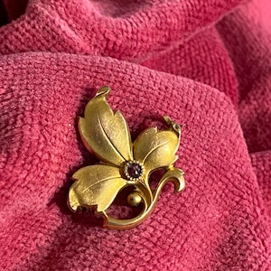 Puede incluir: Un broche floral dorado con una gema púrpura central. El broche presenta diseños detallados de hojas y pétalos, con un elemento de tallo curvo. La joya está sobre un fondo rosa texturizado.