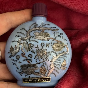 May include: A light blue, round glass bottle with a dark purple cap. The bottle features a map of Devon, England, with silver-colored illustrations and text. The words "BLUE of DEVON" are displayed on a gold label.