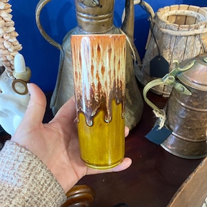 May include: A tall, cylindrical ceramic vase with a dripped glaze effect. The top section is a warm orange-brown, changing to a lighter brown and then a mustard yellow at the base. The vase is held in a hand.