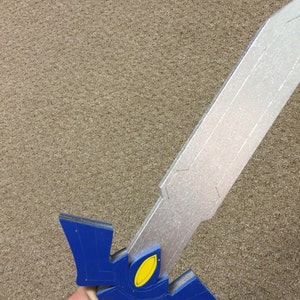 May include: A blue and silver toy sword with a yellow detail. The sword is made of foam and is held in a hand.