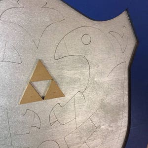 Puede incluir: Un escudo plateado con un borde azul. El escudo presenta un dise&ntilde;o estilizado con una figura de p&aacute;jaro y un emblema triangular marr&oacute;n. El dise&ntilde;o est&aacute; delineado en negro.