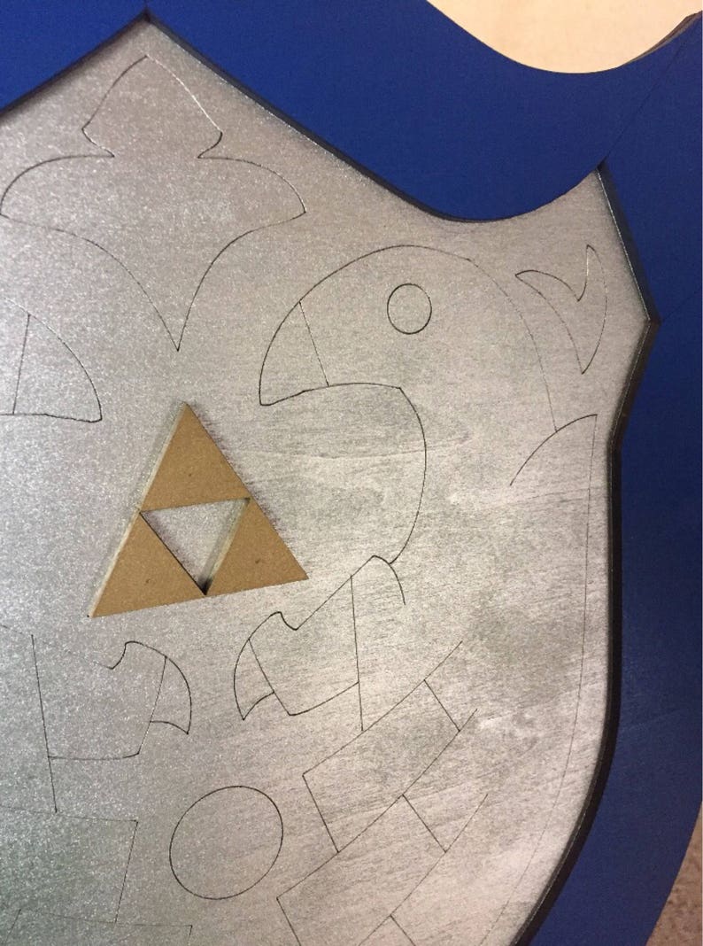 Puede incluir: Un escudo plateado con un borde azul. El escudo presenta un dise&ntilde;o estilizado con una figura de p&aacute;jaro y un emblema triangular marr&oacute;n. El dise&ntilde;o est&aacute; delineado en negro.