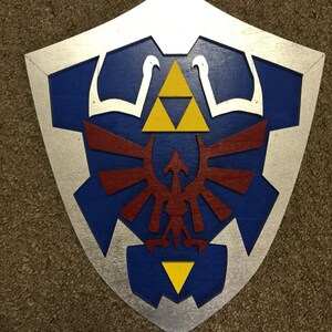 May include: A wooden shield painted in a silver, blue, red, and yellow colour scheme. The shield features a stylised design of a bird with wings spread, a triangle, and a three-pointed star.