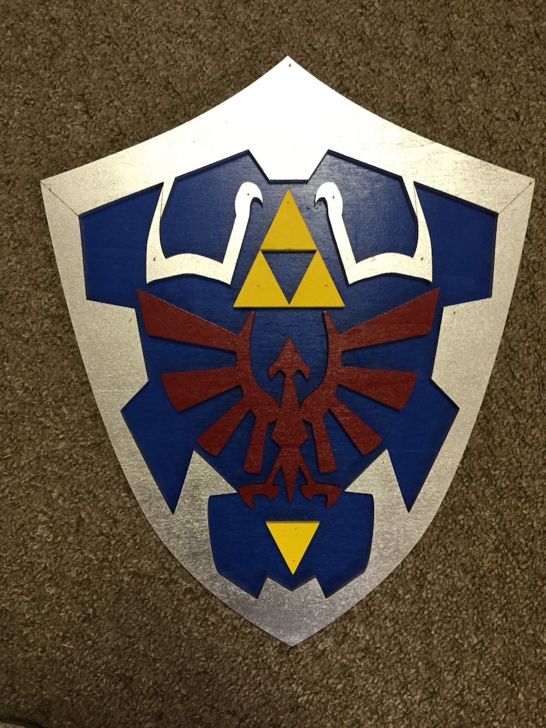 May include: A wooden shield painted in a silver, blue, red, and yellow colour scheme. The shield features a stylised design of a bird with wings spread, a triangle, and a three-pointed star.